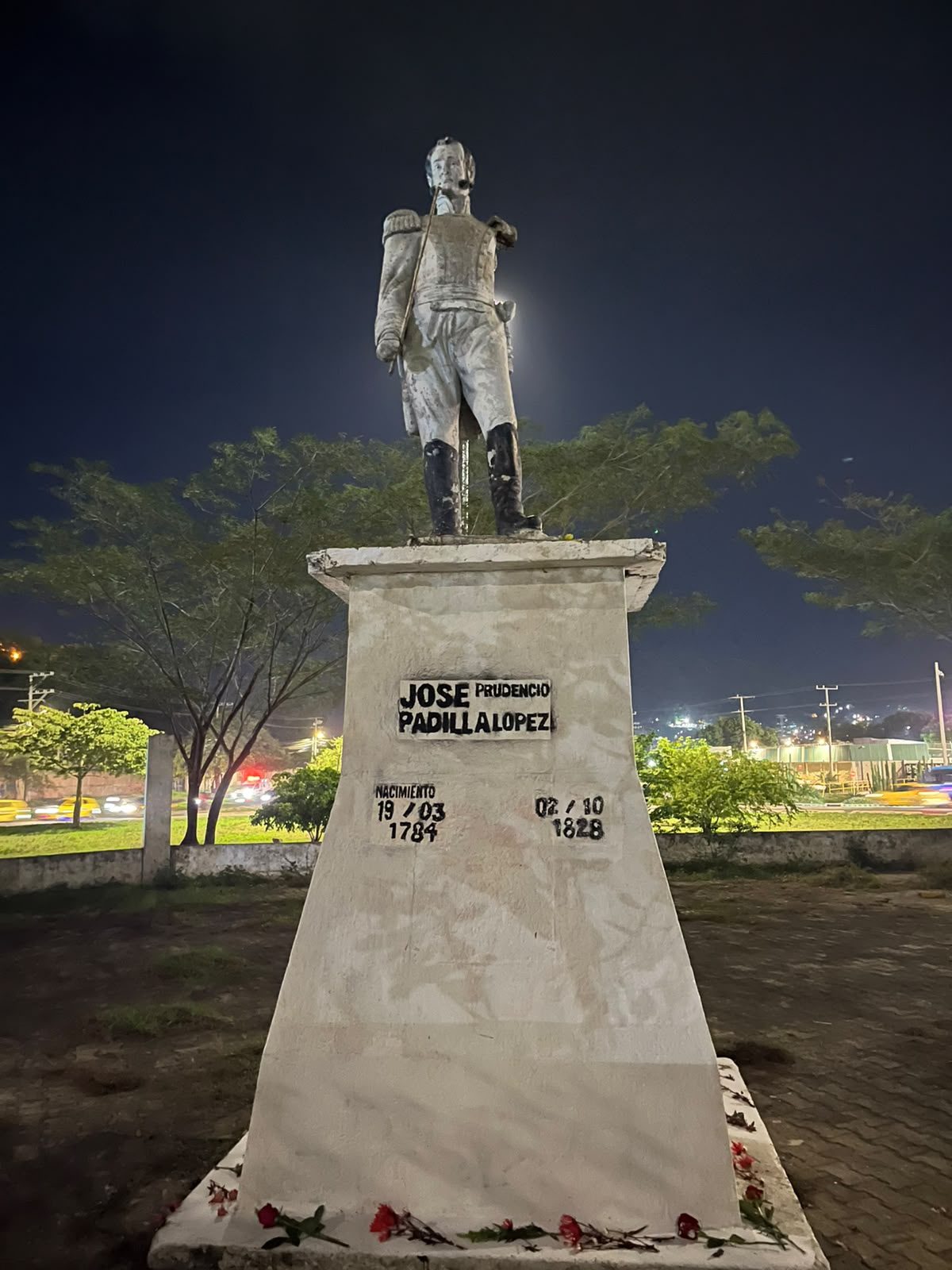 Estatua blanca de José Prudencio Padilla, de noche, sobre un pedestal recién pintado de blanco. En la base, una placa improvisada en pintura negra dice «José Prudencio Padilla López», con las fechas 19/03/1784 y 02/10/1828; al pie, flores rojas.